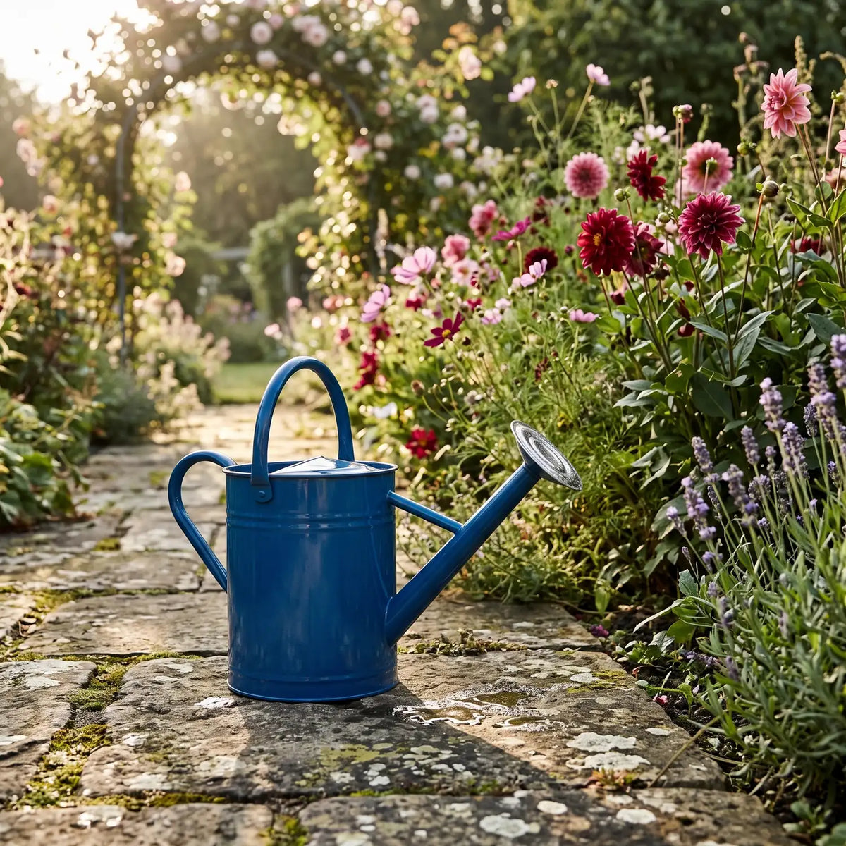Watering Can 8 Litres in blue on a stone path beside a cottage garden border
